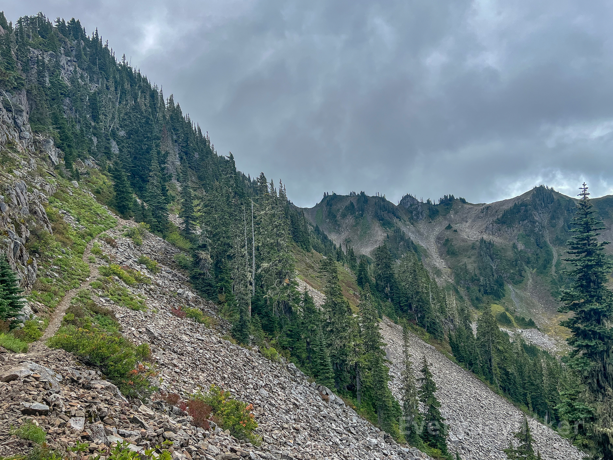 A steep hillside with the trail cutting up the left of frame.  Some patches of trees may be seen hanging on in this heavily sloped scree field.  A jagged ridgeline may be seen in the distance along the middle right under cloudy skies barely above the ridge.