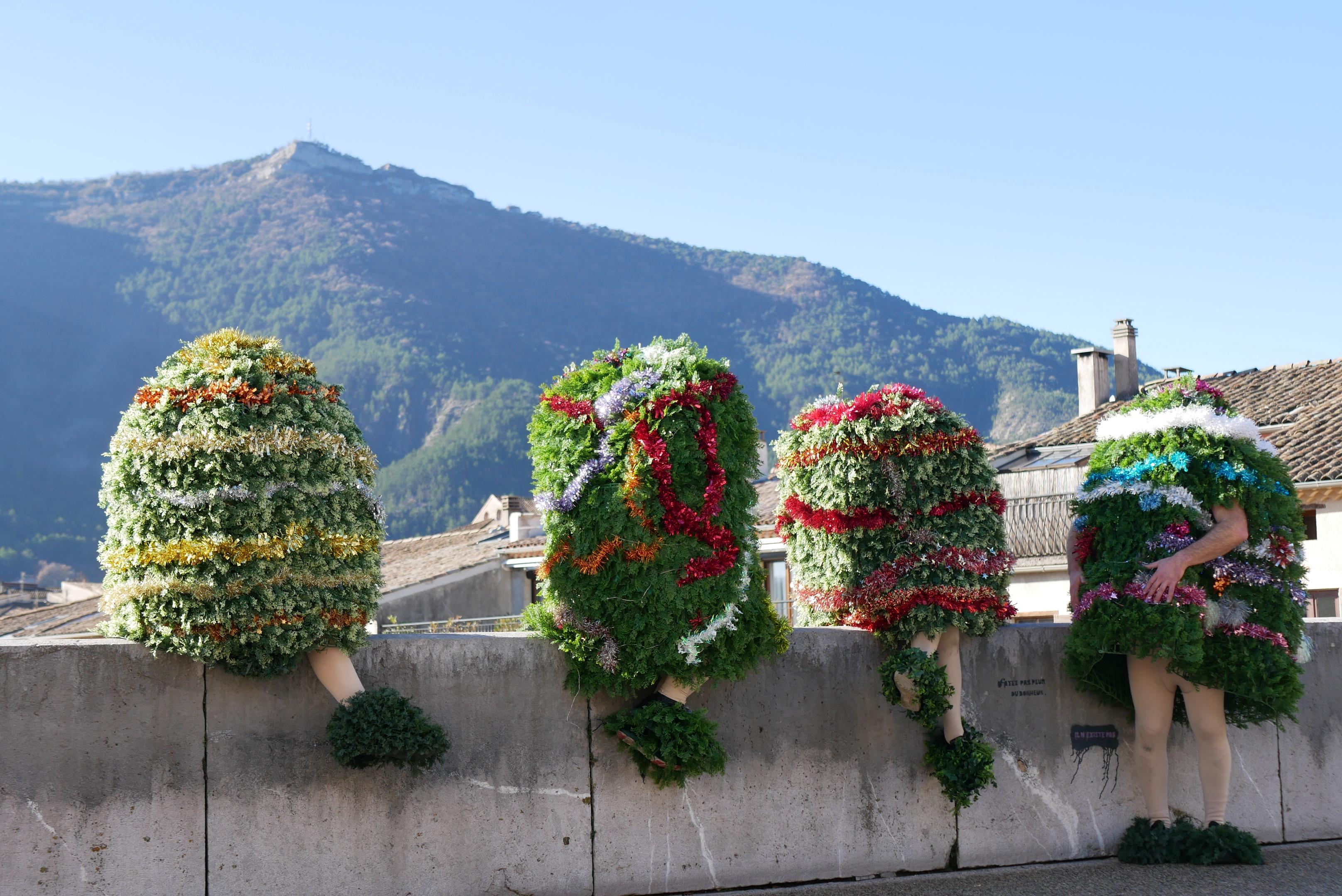 Photographie de quatre personnes déguisées en sapins de Noël, assises au bord d’un pont. On ne voit pas leur tête, seules les jambes (voire les bras) dépassent du « sapin », et ça donne un impression assez bizarre et rigolote. Il y a des toits de maisons et une jolie montagne en arrière-plan.